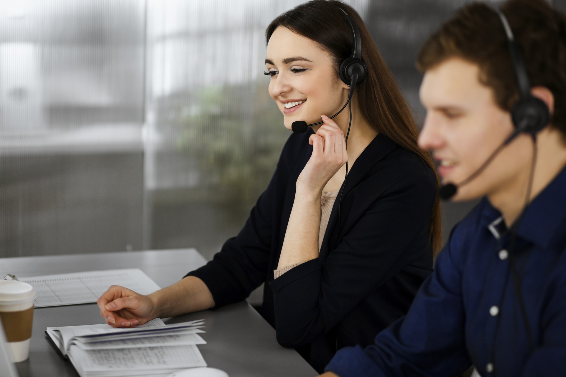 Young beautiful woman casual dressed in headsets is listening to a company's client, while she is sitting at the desk, working together with a male colleague in a modern office. Focus on woman. Call center operators at work